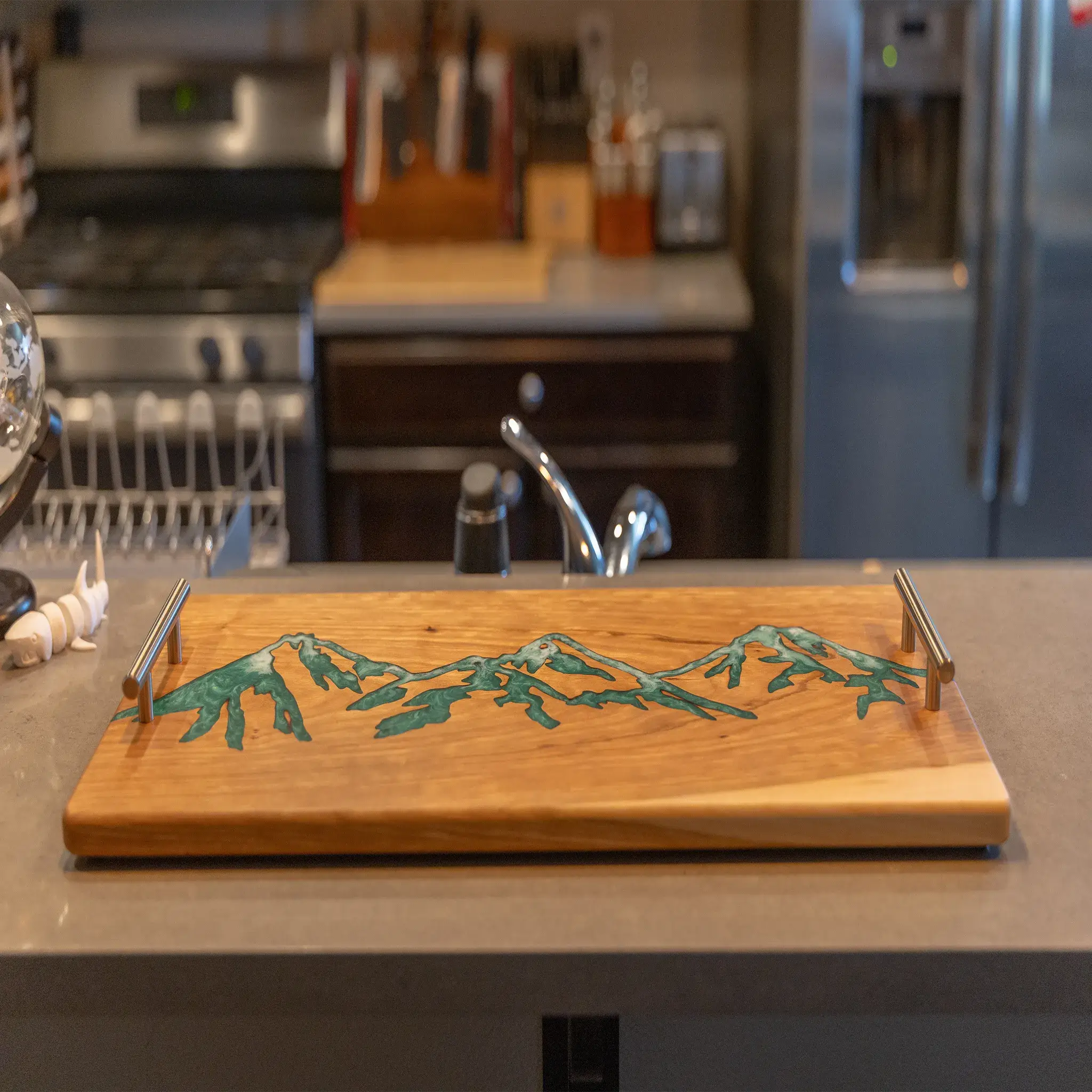 Handcrafted cherry wood charcuterie board featuring a green epoxy mountain inlay displayed on a kitchen countertop.