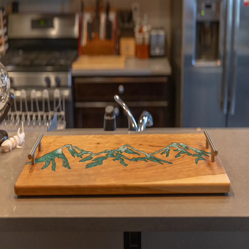 Handcrafted cherry wood charcuterie board featuring a green epoxy mountain inlay displayed on a kitchen countertop.